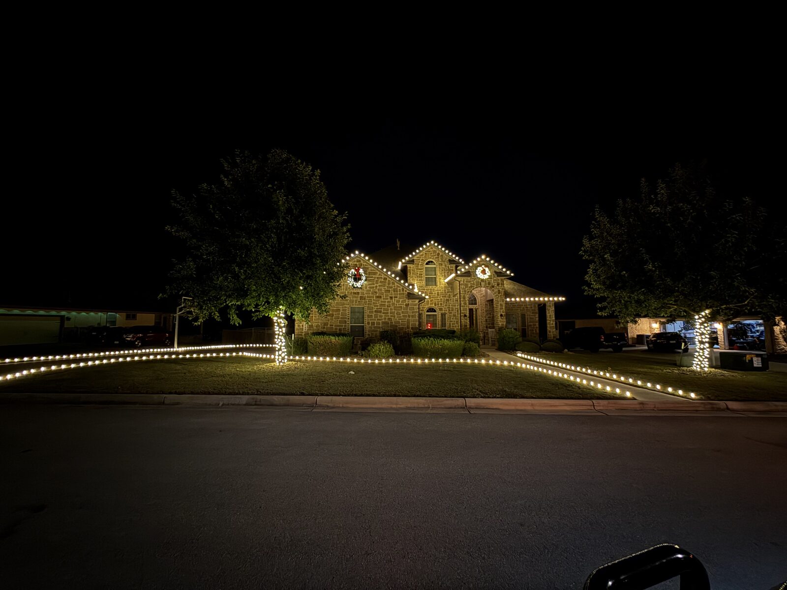 Two-story stone home with lit gables, wreaths, and wrapped trees